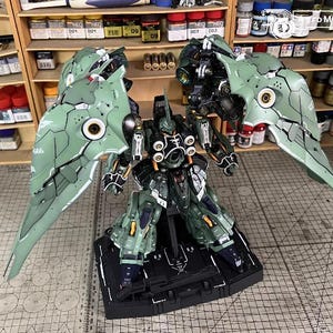May include: A detailed model of a mech robot with large, articulated wings. The robot is primarily green and black, with intricate detailing and a complex design. It stands on a black base, with a grid-patterned surface in the background.