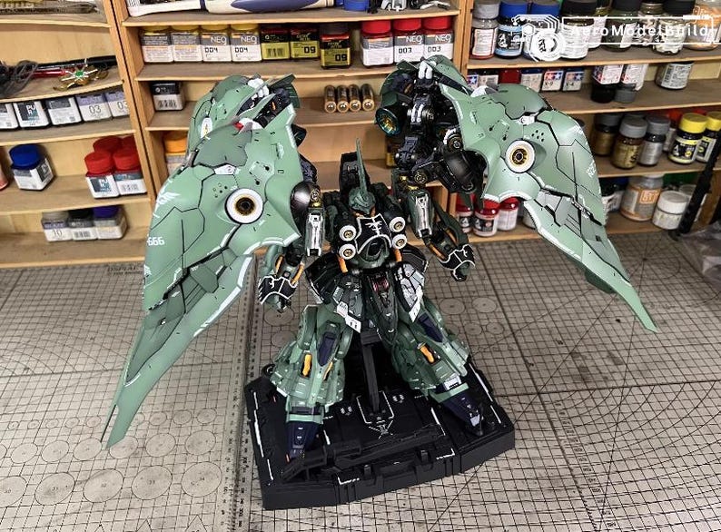 May include: A detailed model of a mech robot with large, articulated wings. The robot is primarily green and black, with intricate detailing and a complex design. It stands on a black base, with a grid-patterned surface in the background.