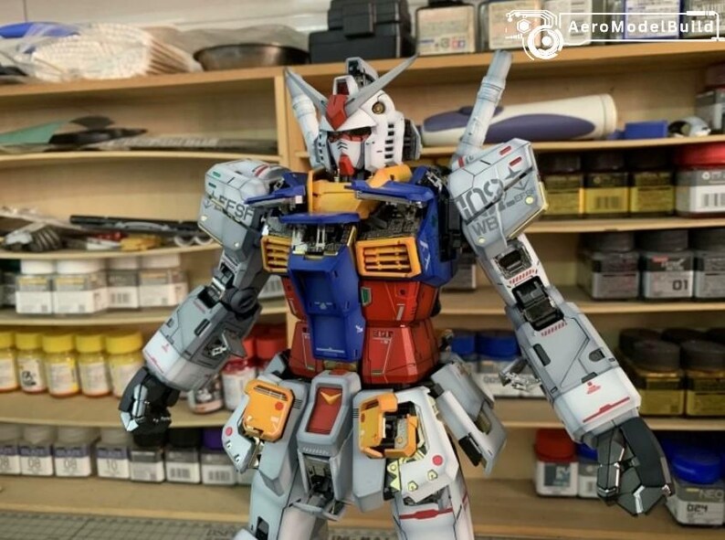 K&ouml;nnte beinhalten: Ein detailliertes Modell eines Gundam-Roboters mit einem Farbschema aus Wei&szlig;, Blau, Rot und Gelb. Der Roboter hat bewegliche Gliedma&szlig;en und komplizierte Linien. Das Modell wird vor Regalen mit Malutensilien ausgestellt.