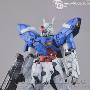 May include: A detailed model of a Gundam robot, with a colour scheme of blue, white, red, and gold. The robot has intricate panel lines and mechanical details, with a large weapon attached to its side. The model is posed against a neutral background.