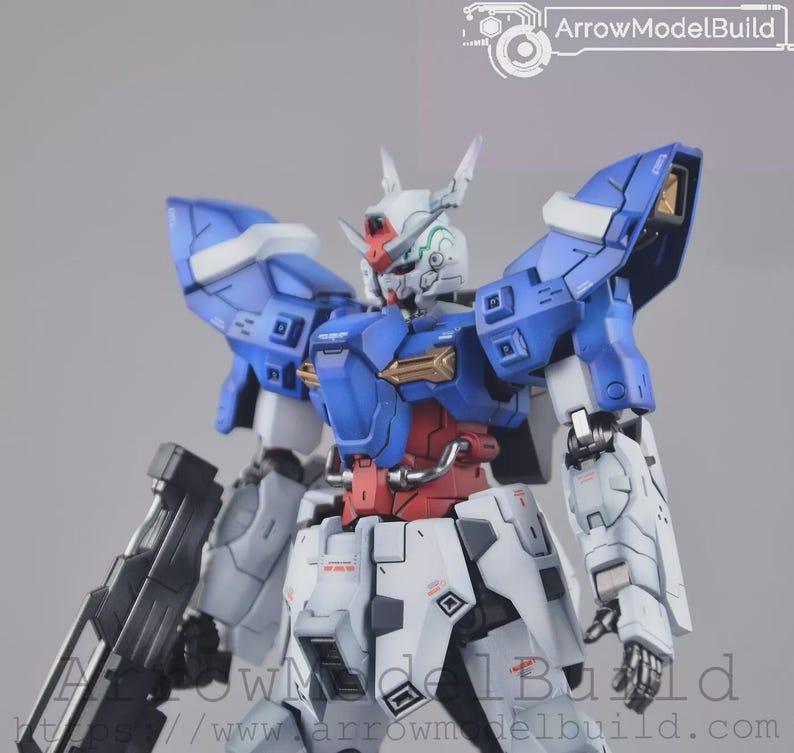 May include: A detailed model of a Gundam robot, with a colour scheme of blue, white, red, and gold. The robot has intricate panel lines and mechanical details, with a large weapon attached to its side. The model is posed against a neutral background.