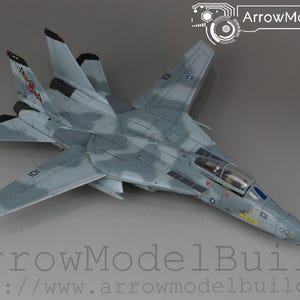 May include: A detailed model aircraft in grey with camouflage patterns. The model has wings, a cockpit, and tail fins. The text "ArrowModelBuild" and a website address are visible. The model is a replica of a military aircraft.