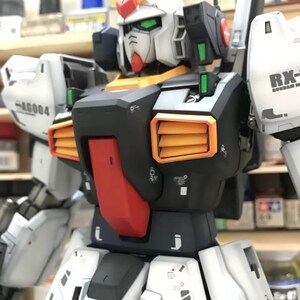 Op de afbeelding: Een gedetailleerd model van een Gundam-robot, voornamelijk wit, zwart en oranje. De robot heeft een opvallende kop met groene ogen en gele en rode accenten. Het model toont fijne details en het "AeroModelBuild" logo.
