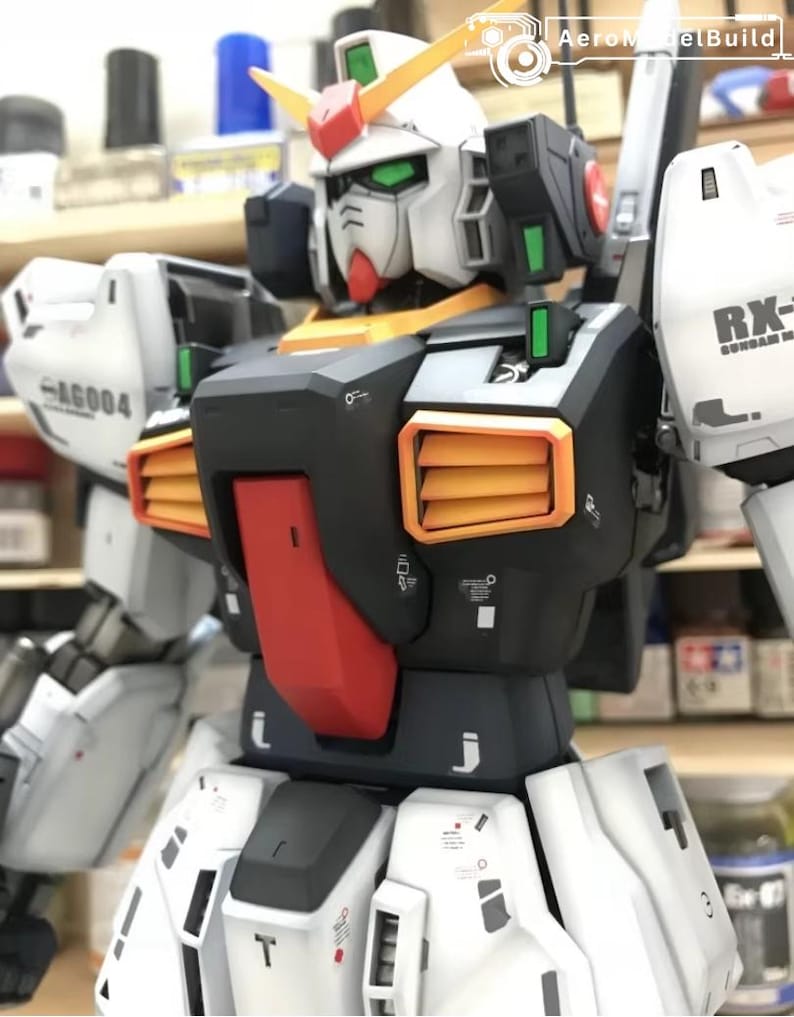 Op de afbeelding: Een gedetailleerd model van een Gundam-robot, voornamelijk wit, zwart en oranje. De robot heeft een opvallende kop met groene ogen en gele en rode accenten. Het model toont fijne details en het "AeroModelBuild" logo.