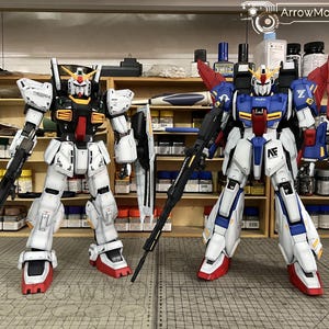 May include: Two detailed Gundam model kits. The model on the left is primarily white with black and red accents, holding a rifle and shield. The model on the right is blue, white, and red, also with a rifle. Both are standing upright.