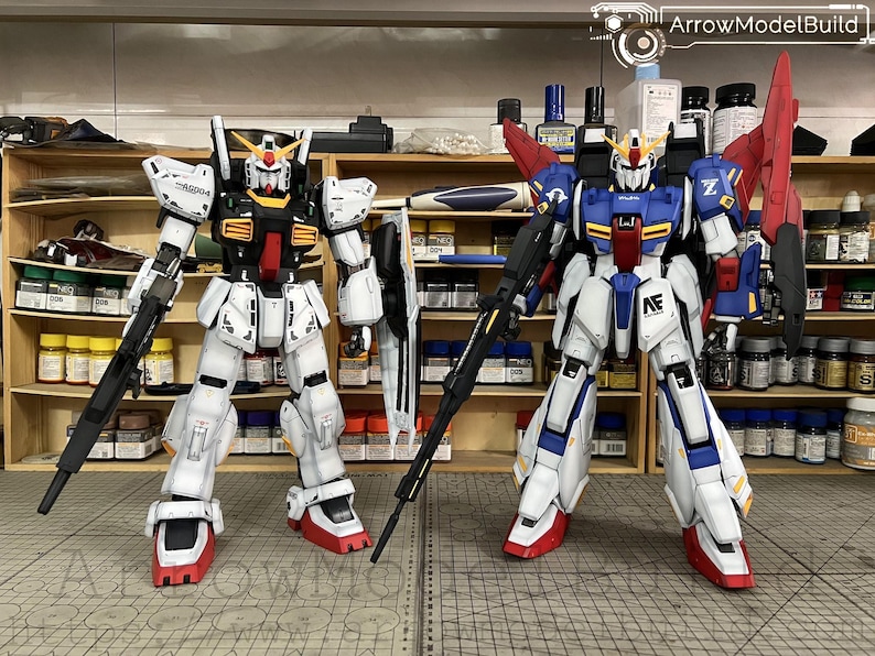 May include: Two detailed Gundam model kits. The model on the left is primarily white with black and red accents, holding a rifle and shield. The model on the right is blue, white, and red, also with a rifle. Both are standing upright.
