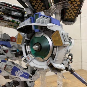May include: Detailed view of a white, blue, and gray model robot with a large circular component featuring a green center and yellow accents. The model is supported by clear stands. The text "ArrowModelBuild" is visible.