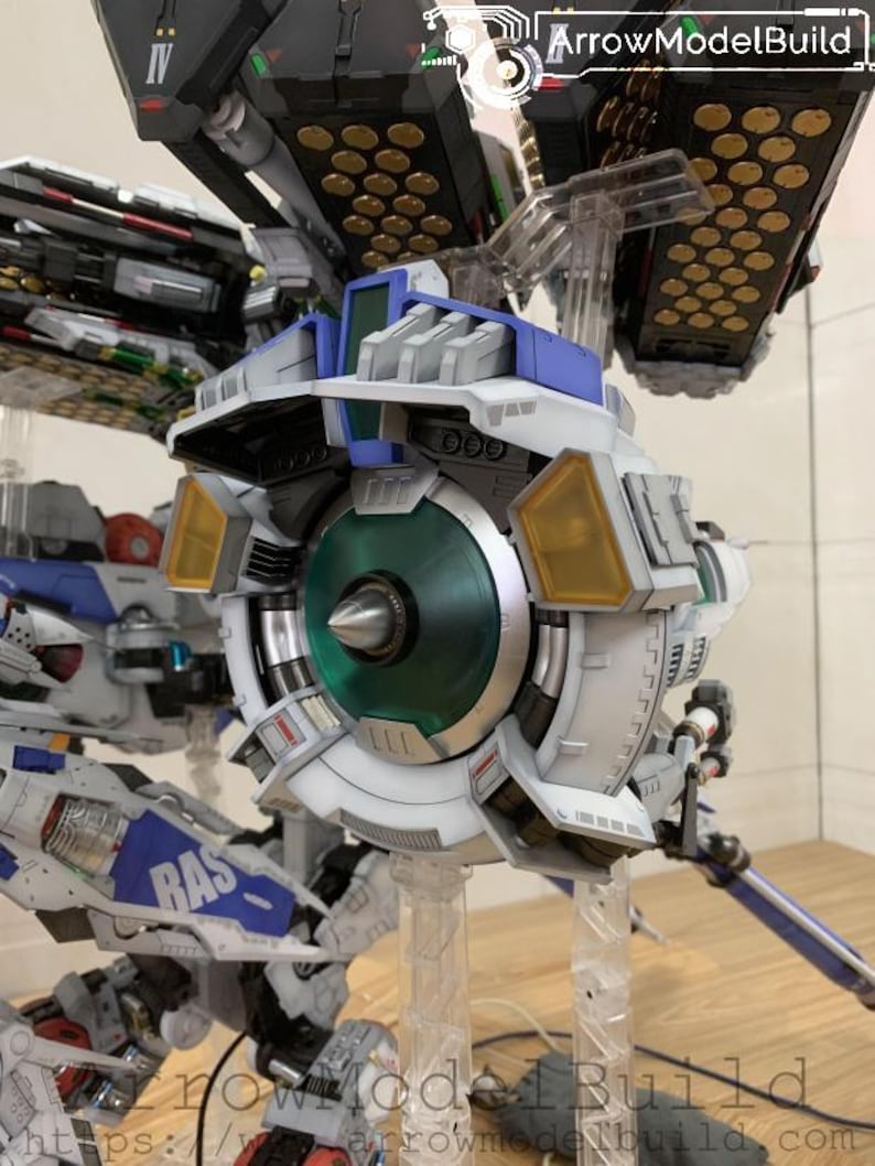 May include: Detailed view of a white, blue, and gray model robot with a large circular component featuring a green center and yellow accents. The model is supported by clear stands. The text "ArrowModelBuild" is visible.