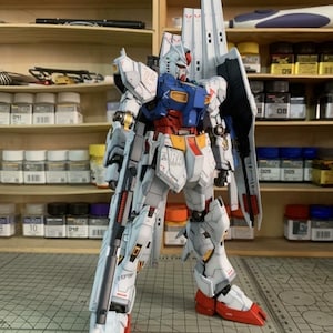 K&ouml;nnte beinhalten: Ein detailliertes Modell eines wei&szlig;en, blauen, roten und gelben Mecha-Roboters mit einem gro&szlig;en Gewehr. Das Modell hat komplizierte Linien und Markierungen, darunter "EFSF" und "VRA". Der Roboter steht auf einer Rasteroberfl&auml;che, mit Regalen voller Modellbauzubeh&ouml;r im Hintergrund.