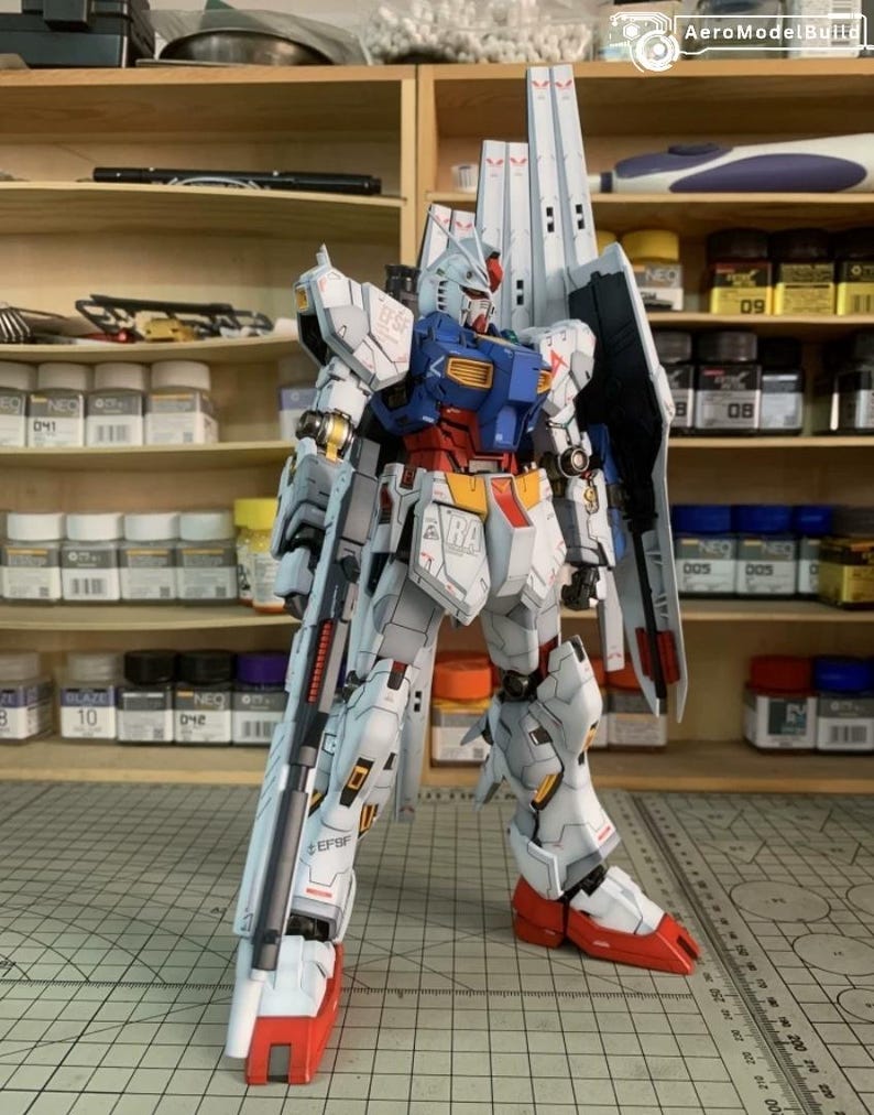 K&ouml;nnte beinhalten: Ein detailliertes Modell eines wei&szlig;en, blauen, roten und gelben Mecha-Roboters mit einem gro&szlig;en Gewehr. Das Modell hat komplizierte Linien und Markierungen, darunter "EFSF" und "VRA". Der Roboter steht auf einer Rasteroberfl&auml;che, mit Regalen voller Modellbauzubeh&ouml;r im Hintergrund.