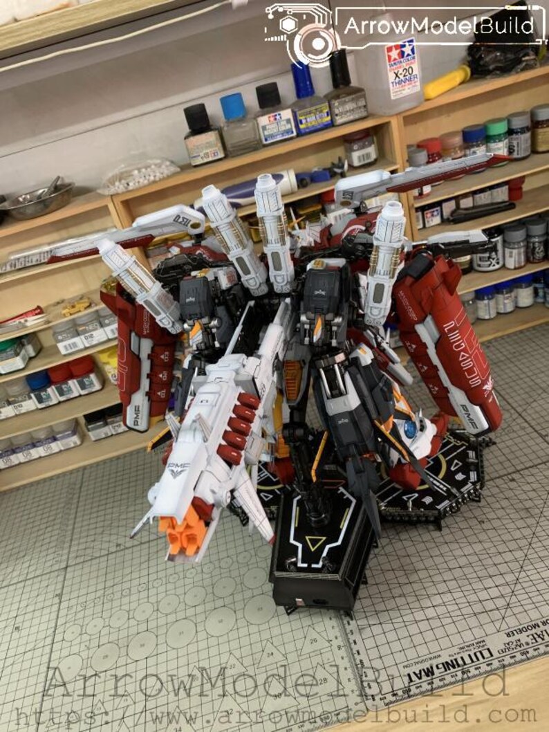 Puede incluir: Un kit de modelo detallado de un robot mec&aacute;nico, con un dise&ntilde;o complejo con acentos rojos, blancos, negros y naranjas. El modelo tiene m&uacute;ltiples partes articuladas y se muestra en un soporte negro. El texto "ArrowModelBuild" es visible.