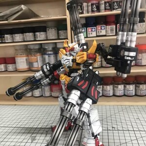 K&ouml;nnte beinhalten: Ein detailliertes Modell eines Roboters mit mehreren Kanonen. Der Roboter ist haupts&auml;chlich wei&szlig;, gelb und rot, mit schwarzen Akzenten. Das Modell ist mit ausgestreckten Armen posiert, um das komplizierte Design und die Waffen zu pr&auml;sentieren. Der Hintergrund umfasst Regale mit verschiedenen Farbt&ouml;pfen.