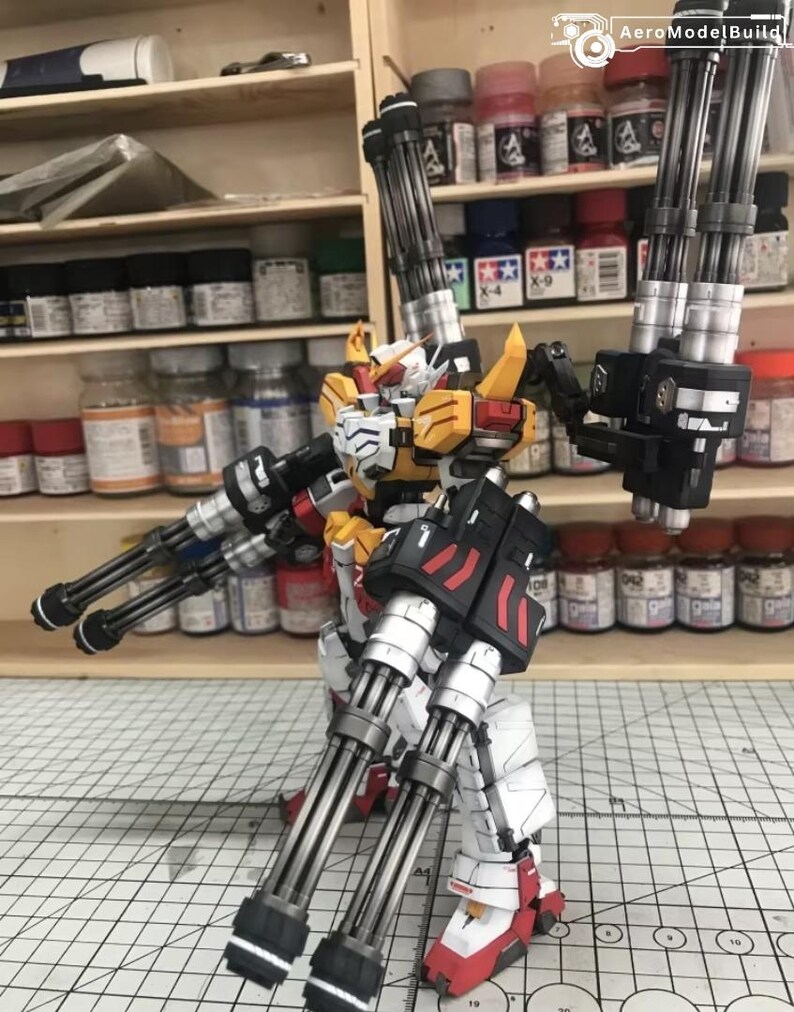 K&ouml;nnte beinhalten: Ein detailliertes Modell eines Roboters mit mehreren Kanonen. Der Roboter ist haupts&auml;chlich wei&szlig;, gelb und rot, mit schwarzen Akzenten. Das Modell ist mit ausgestreckten Armen posiert, um das komplizierte Design und die Waffen zu pr&auml;sentieren. Der Hintergrund umfasst Regale mit verschiedenen Farbt&ouml;pfen.