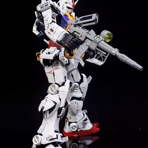 Op de afbeelding: Gedetailleerd model van een witte en rode robot met een groot grijs geweer. De robot heeft een rode en gele kop met een witte romp en benen. De robot staat op een zwart oppervlak.