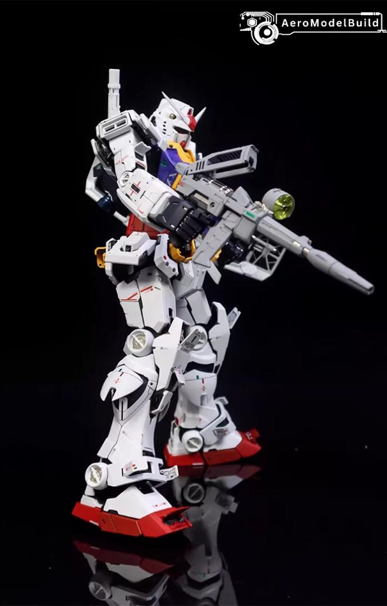 Op de afbeelding: Gedetailleerd model van een witte en rode robot met een groot grijs geweer. De robot heeft een rode en gele kop met een witte romp en benen. De robot staat op een zwart oppervlak.