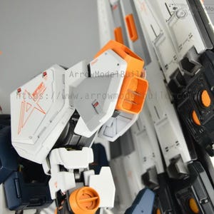 May include: Close-up of a detailed white, orange, and navy blue model kit. The model features intricate panel lines, orange accents, and a complex mechanical design. The text "ArrowModelBuild" is visible.
