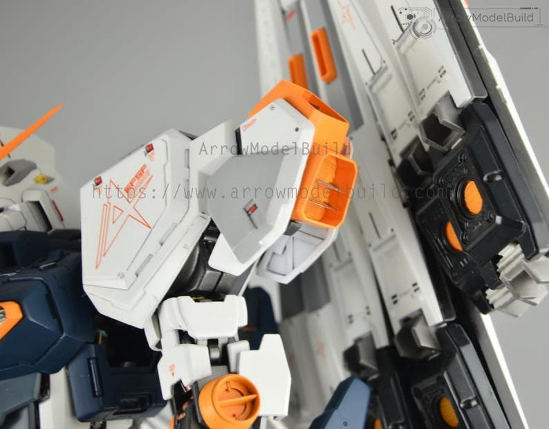 May include: Close-up of a detailed white, orange, and navy blue model kit. The model features intricate panel lines, orange accents, and a complex mechanical design. The text "ArrowModelBuild" is visible.