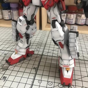 K&ouml;nnte beinhalten: Nahaufnahme der Beine eines detaillierten, beweglichen Modellroboters. Die Beine sind haupts&auml;chlich wei&szlig; mit roten und gelben Akzenten. Das Modell steht auf einer Rasteroberfl&auml;che.