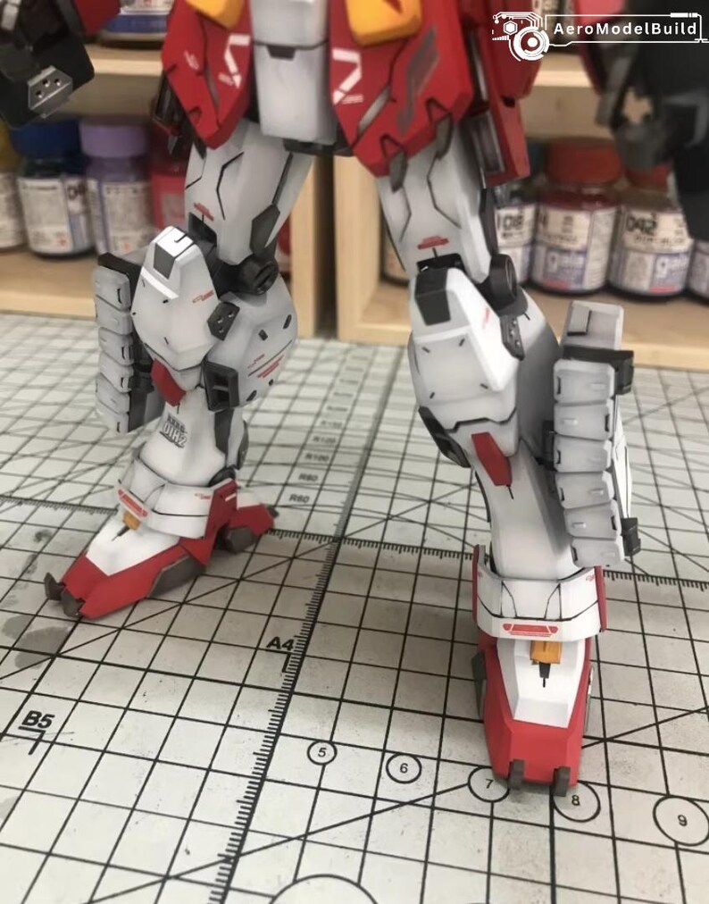 K&ouml;nnte beinhalten: Nahaufnahme der Beine eines detaillierten, beweglichen Modellroboters. Die Beine sind haupts&auml;chlich wei&szlig; mit roten und gelben Akzenten. Das Modell steht auf einer Rasteroberfl&auml;che.