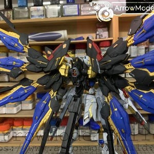 Puede incluir: Kit de modelo detallado de un robot mecha con grandes alas estilizadas. El robot presenta una combinaci&oacute;n de colores azul, dorado, negro y gris. Las alas son predominantemente azules con detalles dorados. El modelo se exhibe sobre un fondo de estantes con varios suministros de hobby.