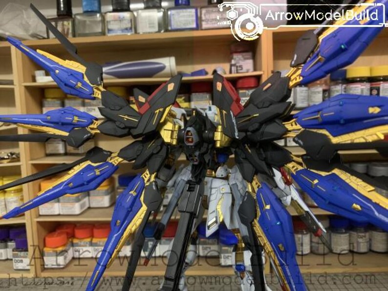 Puede incluir: Kit de modelo detallado de un robot mecha con grandes alas estilizadas. El robot presenta una combinaci&oacute;n de colores azul, dorado, negro y gris. Las alas son predominantemente azules con detalles dorados. El modelo se exhibe sobre un fondo de estantes con varios suministros de hobby.