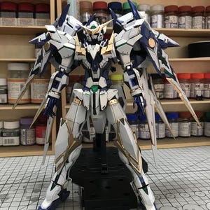 Puede incluir: Un modelo detallado de un robot mecha blanco, azul y dorado con armadura intrincada y bordes afilados. El robot está sobre una base negra, con múltiples alas y cuchillas. El texto "ArrowModelBuild" es visible en la esquina superior derecha.