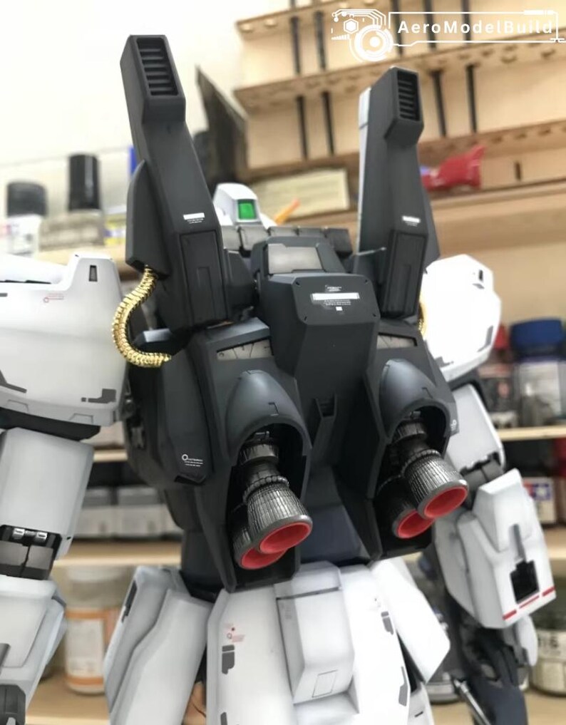 Op de afbeelding: Gedetailleerd model van een witte en zwarte robot met rode accenten op de stuwraketten. De robot heeft een complex ontwerp met meerdere componenten, waaronder grote zwarte boosters en goudkleurige buizen. Het AeroModelBuild-logo is zichtbaar.