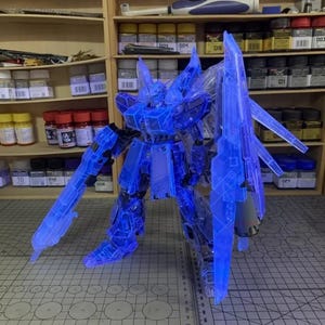 May include: A transparent blue model robot with articulated limbs and weapons, including a large shield. The model is lit up, revealing its internal components. The robot stands on a grid surface, with shelves of model supplies behind it.