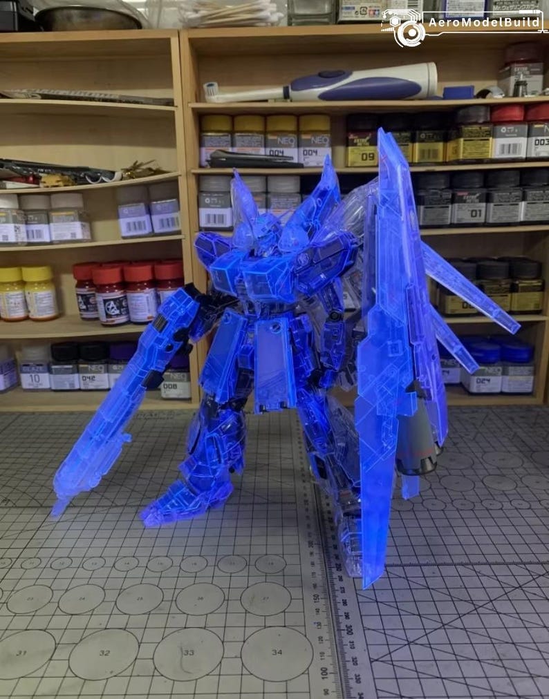 May include: A transparent blue model robot with articulated limbs and weapons, including a large shield. The model is lit up, revealing its internal components. The robot stands on a grid surface, with shelves of model supplies behind it.