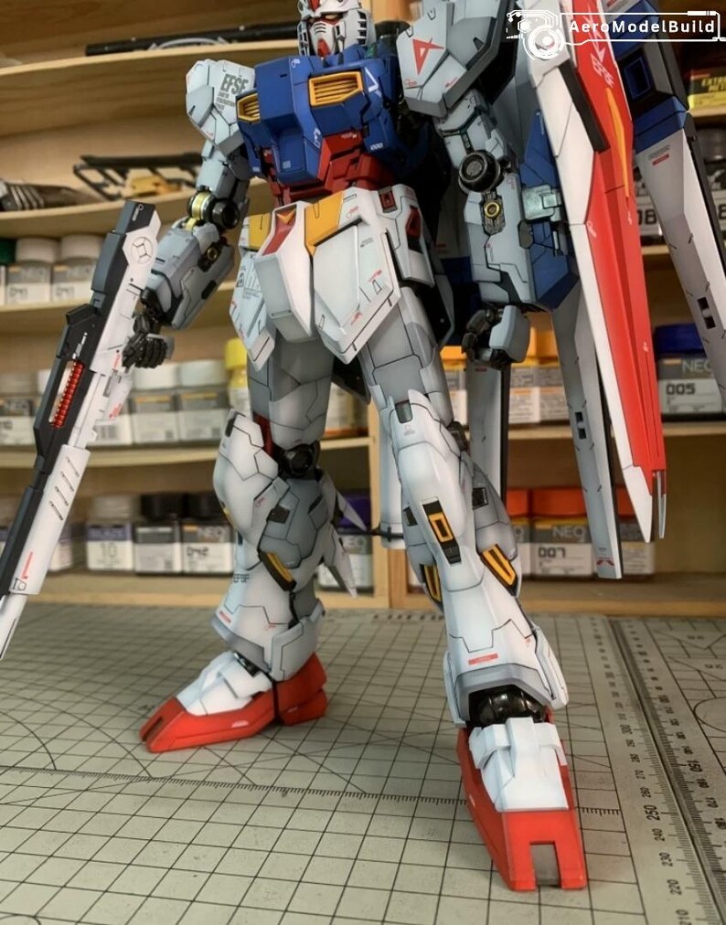 K&ouml;nnte beinhalten: Detailliertes Modell eines Roboters, haupts&auml;chlich wei&szlig;, blau, rot und grau. Der Roboter h&auml;lt eine gro&szlig;e Waffe und hat einen Schild auf dem R&uuml;cken. Das Modell steht auf einer Rasteroberfl&auml;che, mit Regalen und Zubeh&ouml;r im Hintergrund.