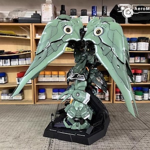 May include: A detailed model of a green and black mech with large, wing-like structures. The model has intricate paneling and circular details, with a black base. The background shows shelves with paint bottles and tools, suggesting a hobby or model-building setting.