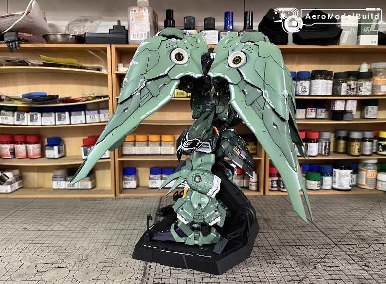 May include: A detailed model of a green and black mech with large, wing-like structures. The model has intricate paneling and circular details, with a black base. The background shows shelves with paint bottles and tools, suggesting a hobby or model-building setting.
