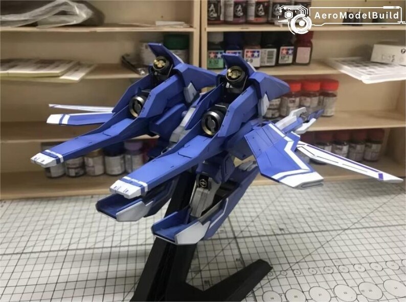 May include: A blue and white model airplane with a detailed design. The model has a sleek, aerodynamic shape with wings and a tail. The model is on a black stand. The AeroModelBuild logo is in the top right corner.