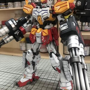 K&ouml;nnte beinhalten: Detailliertes Modell eines Gundam-Roboters, haupts&auml;chlich wei&szlig; mit roten, gelben und schwarzen Akzenten. Der Roboter ist mit gro&szlig;en Waffen mit mehreren L&auml;ufen an jedem Arm ausgestattet. Das Modell steht auf einer Schneidematte, was auf ein Hobbyprojekt hindeutet.