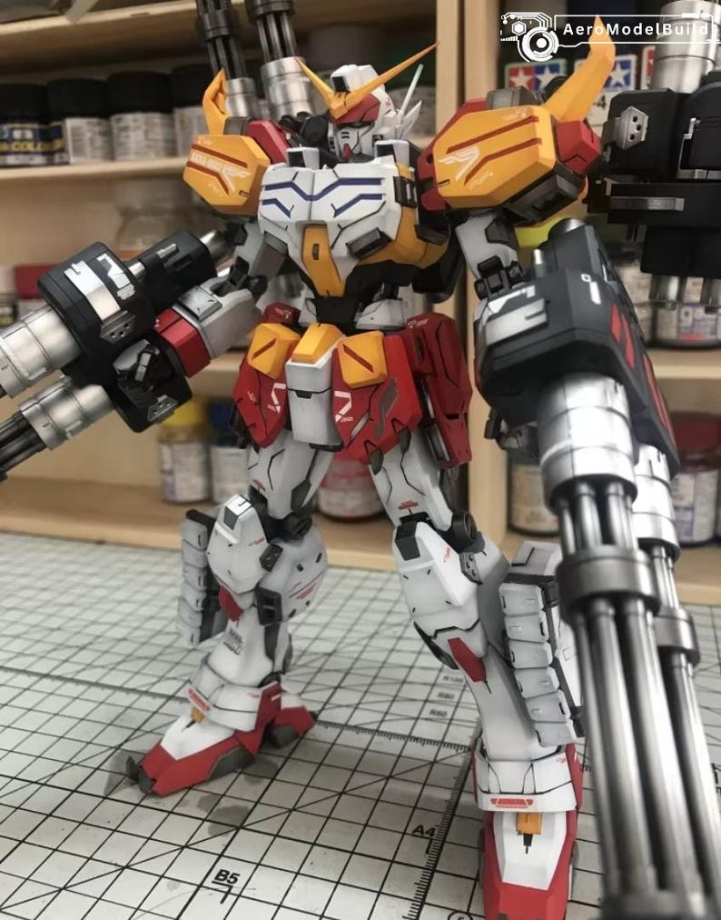 K&ouml;nnte beinhalten: Detailliertes Modell eines Gundam-Roboters, haupts&auml;chlich wei&szlig; mit roten, gelben und schwarzen Akzenten. Der Roboter ist mit gro&szlig;en Waffen mit mehreren L&auml;ufen an jedem Arm ausgestattet. Das Modell steht auf einer Schneidematte, was auf ein Hobbyprojekt hindeutet.