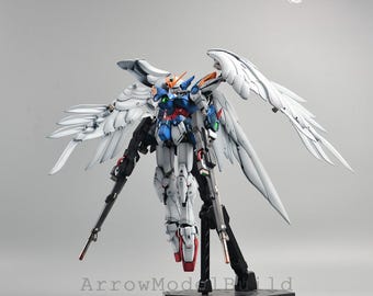 Könnte beinhalten: Detailliertes Modell eines Gundam-Roboters mit großen weißen Flügeln, blauer, roter und weißer Rüstung und zwei schwarzen Gewehren. Das Modell steht auf einem schwarzen Ständer. Der Text "ArrowModelBuild" ist unten sichtbar.