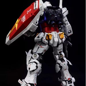 Op de afbeelding: Gedetailleerd model van een Gundam-robot, voornamelijk wit met rode, blauwe en gele accenten. De robot heeft een schild met een sterembleem en tekst. Het model staat tegen een zwarte achtergrond.