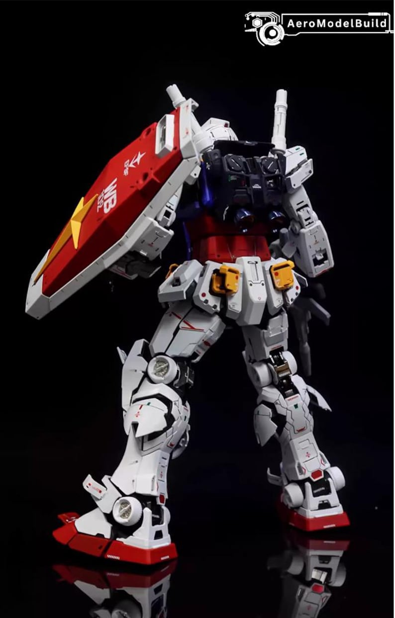 Op de afbeelding: Gedetailleerd model van een Gundam-robot, voornamelijk wit met rode, blauwe en gele accenten. De robot heeft een schild met een sterembleem en tekst. Het model staat tegen een zwarte achtergrond.