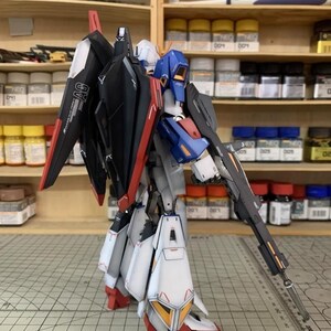 May include: A detailed model robot in black, red, white, blue, and yellow. The robot is holding a long gun and has large wings. The model is on a cutting mat with a grid pattern. The background includes shelves with paint bottles.