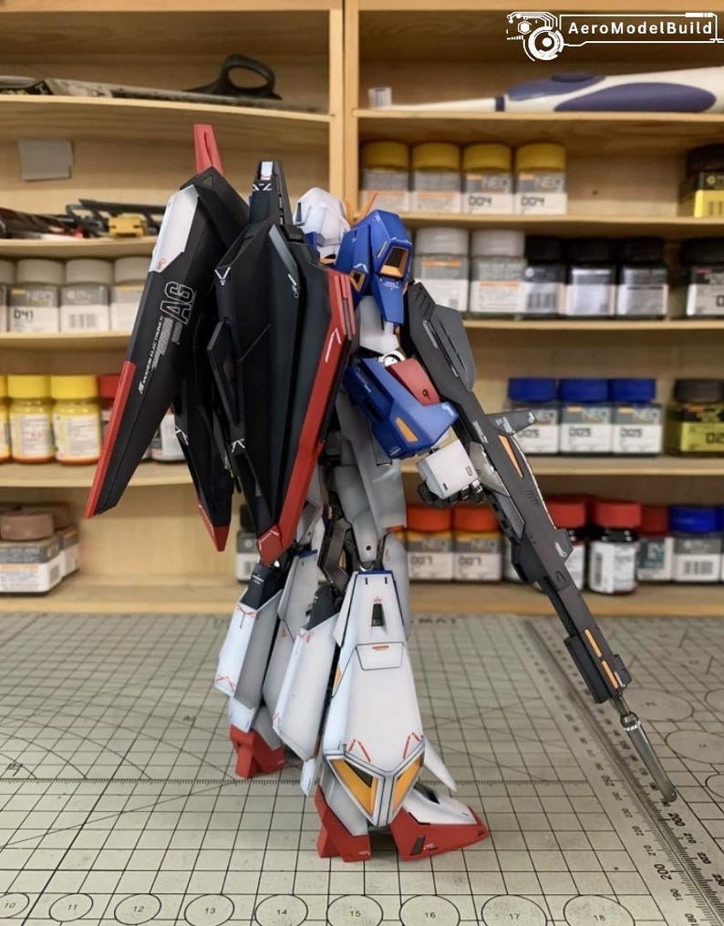 May include: A detailed model robot in black, red, white, blue, and yellow. The robot is holding a long gun and has large wings. The model is on a cutting mat with a grid pattern. The background includes shelves with paint bottles.