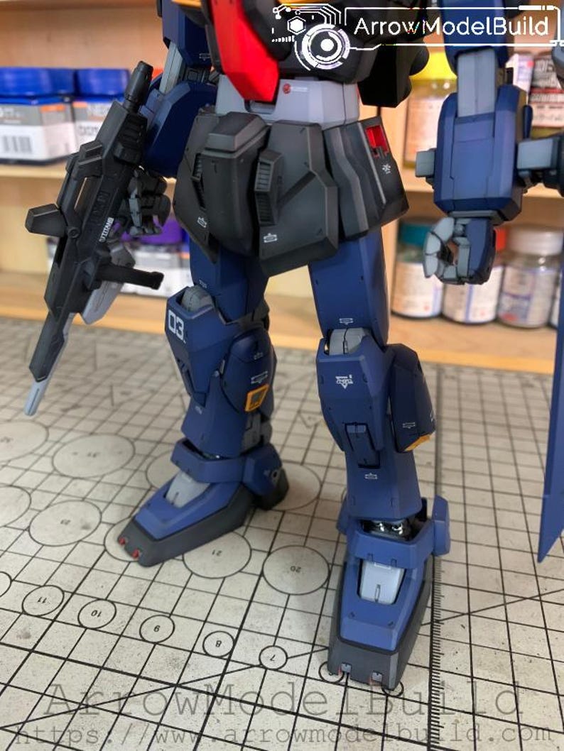 May include: A detailed, partially assembled model robot with a dark blue and black color scheme. The robot has a gun, and the text "03" is visible on the leg. The image also includes the text "Arrow ModelBuild" and a website address.