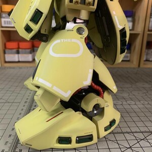 May include: Close-up of a light yellow model robot leg with white accents and green rectangular details. The model has red wiring and the word "THE" is visible. The image is from ArrowModelBuild.