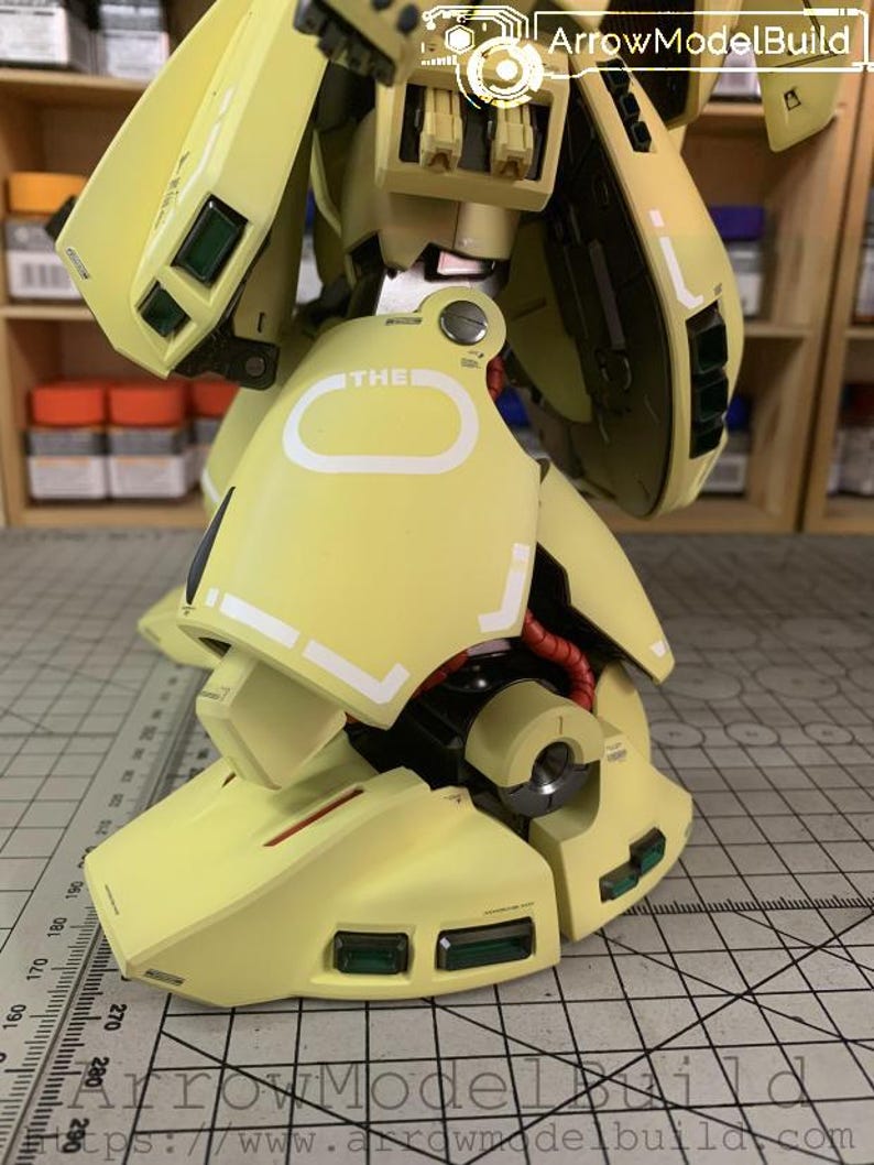 May include: Close-up of a light yellow model robot leg with white accents and green rectangular details. The model has red wiring and the word "THE" is visible. The image is from ArrowModelBuild.