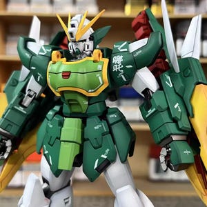 May include: A detailed model of a Gundam robot, predominantly green, white, and yellow. The robot has intricate armour plating, articulated limbs, and a distinctive head design with yellow crests. The model has the text "ArrowModelBuild" and a website address.