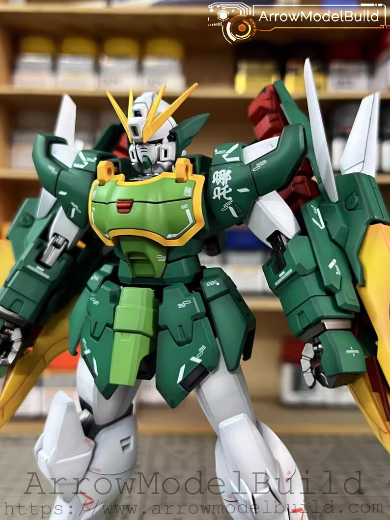 May include: A detailed model of a Gundam robot, predominantly green, white, and yellow. The robot has intricate armour plating, articulated limbs, and a distinctive head design with yellow crests. The model has the text "ArrowModelBuild" and a website address.