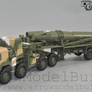 May include: A detailed model of a military truck with a missile launcher. The truck cab is painted in a camouflage pattern of tan, brown, and white. The missile is olive green, mounted on a trailer with multiple wheels.