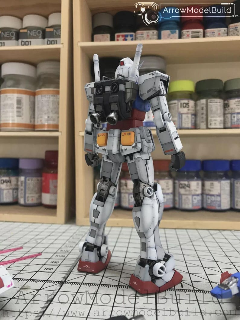 以下が含まれることがあります： 赤、青、黄色のアクセントが付いた、主に白のガンダムロボットの詳細なモデル。モデルは背面から見ており、複雑な機械的詳細が明らかになっています。モデルはグリッド表面に配置されています。