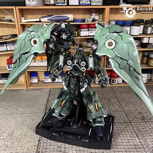 May include: A detailed model of a mech robot with large, articulated wings. The robot is primarily dark green with black and white accents. The wings are a lighter green with intricate paneling. The model is on a black base.