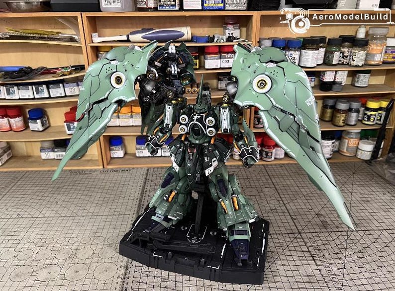 May include: A detailed model of a mech robot with large, articulated wings. The robot is primarily dark green with black and white accents. The wings are a lighter green with intricate paneling. The model is on a black base.