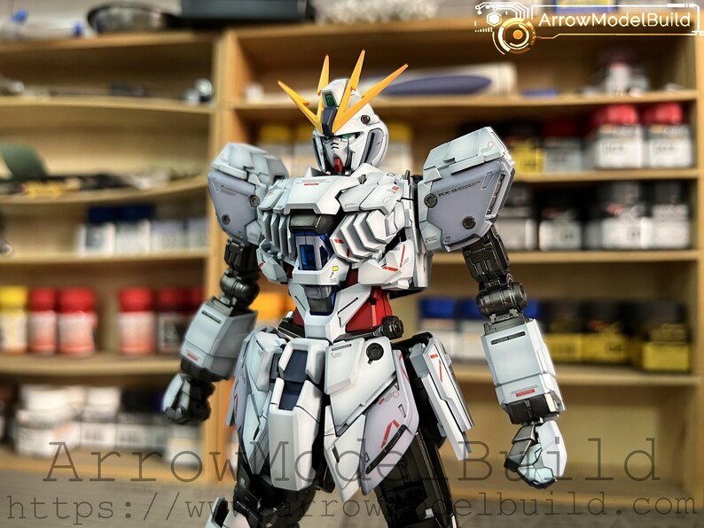 May include: A detailed model of a white, gray, red, and black robot with yellow accents. The robot has a complex design with visible mechanical parts and is posed in a standing position. The background includes shelves with various items.